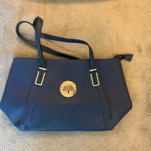 Mörkblå handväska med gulddetaljer mulberry liknande - Snygg mörkblå handväska i slätt läder med axelrem och dekorativt guldfärgat emblem framtill. Väskan har stilrena sömmar och dragkedja upptill. Perfekt för dig som gillar klassisk och trendig design.