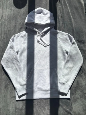 RusseMerch Hoodie grå - Ljusgrå hoodie från Russedress med broderad logga på ärmen. Klassisk modell med huva, dragsnören och stor magficka. Tillverkad i mjukt och bekvämt material, perfekt för chill dagar eller när du vill ha en clean look.