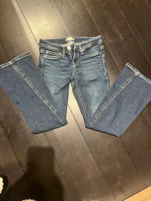 Snygga Blå jeans med vita sömmar! - Säljer ett par blå low rise utsvängda jeans med snygga vita detaljer. Jeansen har dubbla knappar i midjan, klassiska fem fickor och bakfickor med dekorativa detaljer och knappar. Jeansen är från Gina och är i storlek 140. Perfekta för en avslappnad och trendig look. Skriv vid fler frågor!🫶