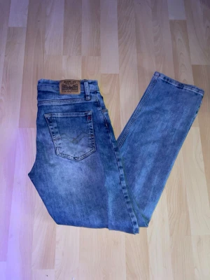 Blå jeans från Replay - Säljer ett par klassiska blå jeans från Replay med snyggt slitna detaljer och raka ben. Jeansen har en bakficka med broderi och ett läderpatch med logotypen på linningen. Materialet är denim och färgen är ljusblå.