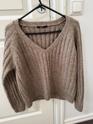 Beige/brun ribbstickad tröja V-ringad - Mysig beige/brun stickad tröja  med V-ringning och ribbstickat mönster. Tröjan har långa ärmar och breda muddar vid ärmslut och nederkant. 