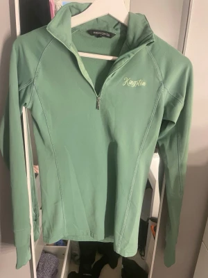 Mintgrön ziptröja från Kingston - Mintgrön långärmad ziptröja från Kingston med halv dragkedja och hög krage. Tröjan har en sportig look och är tillverkad i ett mjukt, stretchigt material som passar perfekt för en aktiv vardag. Diskret logga på bröstet.
