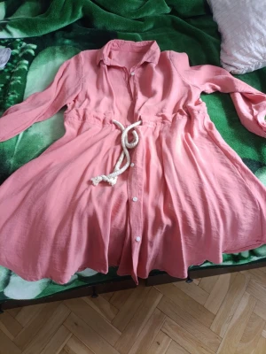 Rosa skjortklänning med knytband - Säljer en söt rosa skjortklänning med långa ärmar och vita knappar framtill. Klänningen har ett vitt repknytband i midjan som ger en avslappnad look. Perfekt för varma dagar och enkel att styla med sneakers eller sandaler.