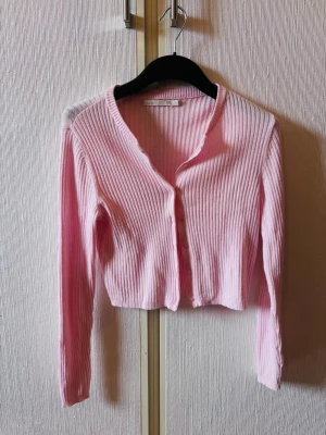 Ljusrosa ribbad kofta - Söt ljusrosa ribbad kofta med lång ärm och knappar framtill. Kortare modell med v-ringad halsringning, perfekt att styla över ett linne eller t-shirt. Materialet är mjukt och stretchigt för en bekväm passform.