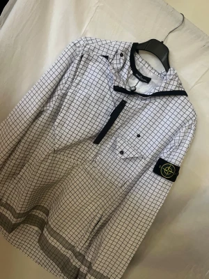 Stone Island rutmönstrad vindjacka - Säljer en vit vindjacka från Stone Island med svart rutmönster och klassisk logopatch på vänster ärm. Jackan har en bröstficka med knappar, dragkedja och detaljer i svart. Perfekt för dig som gillar streetwear och snygga mönster.