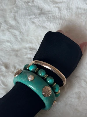 Turkost armband med pärlor och snäckor - Säljer ett coolt turkost armband med stora pärlor och guldfärgade snäckor. Armbandet har en chunky design och är gjort av plast och metall. Perfekt för dig som vill sticka ut och addera färg till din look.
