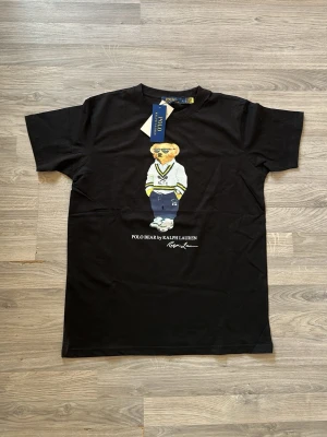 Svart Polo Bear t-shirt från Ralph Lauren - Snygg svart t-shirt från Ralph Lauren med ikoniska Polo Bear-trycket på bröstet. T-shirten har rund hals, korta ärmar och är gjord i mjuk bomull. Perfekt för dig som vill ha en klassisk men lekfull look.