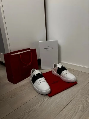 Vltn Skor - Snygga vita sneakers från VLTN med en bred svart läderdetalj över mitten. Klassisk låg modell med rund tå och vita snören. Skorna har en stilren design och är tillverkade i läder. Perfekta för dig som vill ha en exklusiv och trendig look.