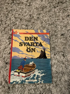 Den svarta ön - Tintins äventyr - Säljer seriealbumet 'Den svarta ön' från Hergés klassiska Tintin-serie. Omslaget är färgglatt med motiv av Tintin och Milou i en båt mot en dramatisk ö. Boken har hårda pärmar och är tryckt på papper. Perfekt för dig som gillar äventyr och serier!