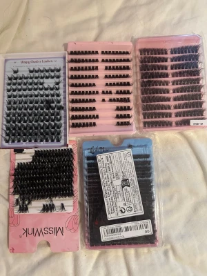 Mixade individuella lösögonfransar - Säljer flera förpackningar med individuella lösögonfransar i olika längder och tjocklekar. Fransarna är svarta och kommer i både naturliga och mer dramatiska stilar. Perfekt för att skapa en personlig look. Förpackningarna är i plast med rosa och lila detaljer.