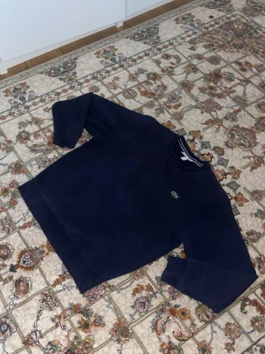 Mörkblå sweatshirt från Lacoste - Klassisk mörkblå sweatshirt från Lacoste med rund halsringning och det ikoniska krokodilmärket broderat på bröstet. Tröjan har ribbade muddar vid ärmslut och nederkant samt en avslappnad passform. Perfekt till jeans eller joggers.