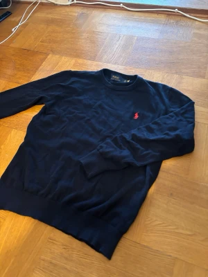  Stickad Polo Ralph Lauren tröja - Mörkblå stickad tröja från Polo Ralph Lauren med rund halsringning och röd broderad logga på bröstet. Tröjan har långa ärmar och ribbade muddar vid ärmslut och nederkant. 
