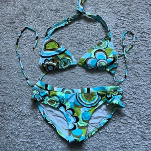 Färgglad blommig bikini med knytband - Säljer en bikini med färgglatt blommönster i blått, grönt, vitt och svart. Överdelen har trekantskupor och knytband i nacken och ryggen. Underdelen har rynkade sidor och matchande mönster. Perfekt för strandhäng eller poolparty!