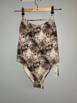 Mönstrad baddräkt med snörning bak - Säljer en snygg baddräkt från Kappahl med trendigt brun-beige mönster och högt skuren modell. Baddräkten har smala axelband och snygg snörning i ryggen som ger en cool detalj. Perfekt för stranden eller poolen!