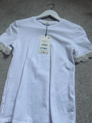 Vit t-shirt med spetsdetalj - Vit t-shirt från zara med korta ärmar och snygga spetsdetaljer vid ärmsluten. Tillverkad i mjuk bomull som känns skön mot huden. Enkel och stilren design som passar perfekt till jeans eller kjol.