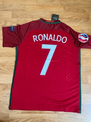 Portugal Ronaldo 7 matchtröja Nike EURO  - Röd Portugal fotbollströja från Nike med nummer 7 och namnet Ronaldo på ryggen. Tröjan har gröna detaljer längs sidorna, officiella märken på ärmarna och Portugals landslagsemblem på bröstet. Perfekt för fotbollsfans som vill sticka ut på planen. Portugal EURO