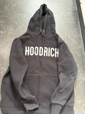 Svart hoodie från Hoodrich  - Svart hoodie från Hoodrich med stor vit logga på bröstet. Klassisk modell med huva och dragsnören samt känguruficka framtill. Tillverkad i mjukt bomullsmaterial som är skönt att bära. Perfekt för en streetwear-look.