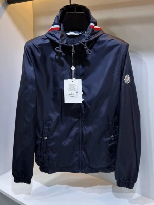 Moncler windbreaker - Stilren mörkblå vindjacka från Moncler med huva och dragkedja framtill. Jackan har röda och vita detaljer vid kragen, två fickor med knapp och Moncler-logga på ärmen. Perfekt för blåsiga dagar och enkel att matcha.