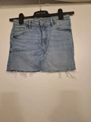 Ljusblå jeanskjol från River Island - Säljer en ljusblå jeanskjol med rå kant nertill och klassisk femficksmodell. Kjolen har normal passform och är tillverkad i bomull. Perfekt för dig som gillar en avslappnad och trendig stil. Hade sytt om den från skinny jeans till en kjol.
