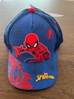 Blå Spiderman keps  - Cool blå keps med Spiderman-motiv . Kepsen har röda detaljer och en stor Spiderman-figur på framsidan samt logotypen på skärmen. Justerbar baktill och tillverkad i bomull. Perfekt för dig som gillar superhjältar och vill sticka ut.