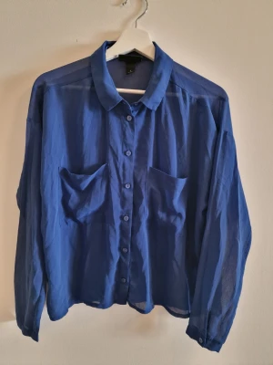 Blå skjorta från Monki med fickor - Snygg blå skjorta från Monki i tunnare material. Skjortan har två bröstfickor, klassisk krage och knäppning framtill. Perfekt för en avslappnad och trendig look.