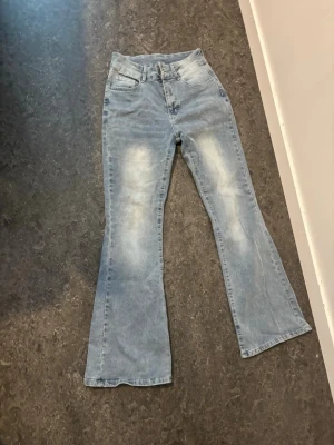 Ljusblå bootcut jeans  - Säljer ett par ljusblå bootcut jeans med snygga slitningar framtill och klassiska fickor bak med knappdetaljer. Jeansen har en utsvängd passform och är tillverkade i denim. Perfekta för en trendig och avslappnad look.