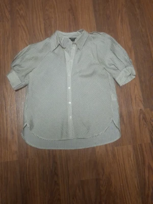 Randig blus med puffärm - Snygg blus med tunna svarta och vita ränder, klassisk krage och puffiga korta ärmar. Knäppning framtill och rundad nederkant ger en trendig look. Perfekt att styla med jeans eller kjol för en cool vardagsstil.