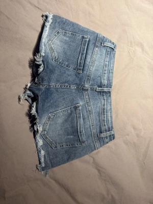 Blå jeansshorts med fransig kant - Säljer ett par klassiska blå jeansshorts med fransig, rå kant och femficksmodell. Shortsen har normal passform och stängs med knapp och dragkedja. Perfekta för sommaren och ger en avslappnad look.
