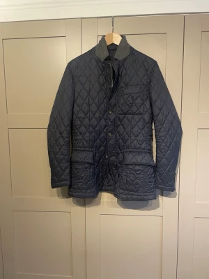 Marinblå quiltad dunjacka - Snygg marinblå dunjacka med quiltat mönster och hög krage i kontrasttyg. Jackan har tre framfickor med lock och knäppning framtill. Passar perfekt för vår och höst. jackan har en klassisk, stilren look.