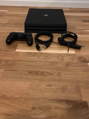 PlayStation 4 Pro med tillbehör - Säljer en svart PlayStation 4 Pro från Sony. Konsolen har en stilren, rektangulär design och är tillverkad i plast. Medföljer en svart DualShock 4-kontroll, HDMI-kabel och strömkabel. Perfekt för gaming och streaming.