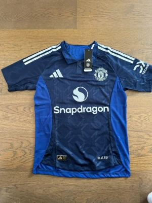 Manchester United matchtröja blå - Snygg blå Manchester United matchtröja från Adidas med vita detaljer och tryck. Tröjan har korta ärmar, krage och logga på bröstet samt sponsortryck. På ryggen står det 'Cunha 10' i vitt. Perfekt för fotbollsfans!
