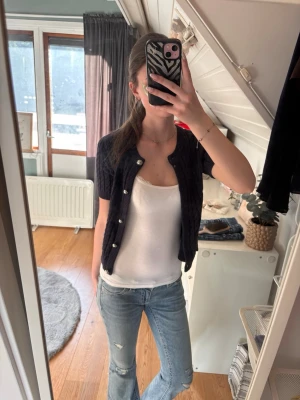 Svart kabelstickad kofta med pärlknappar - Kortärmad mörkblå kabelstickad kofta med snygga silverknappar framtill. Koftan har rund halsringning och sitter figurnära. Perfekt att bära öppen eller stängd över ett linne eller t-shirt.