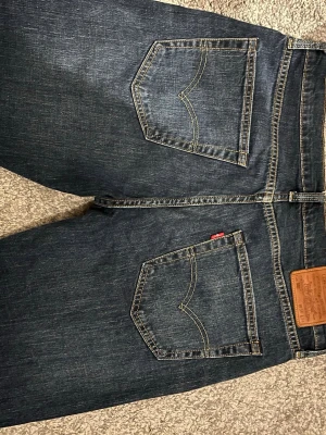 Mörkblå Levi's jeans med raka ben - Snygga mörkblå jeans från Levi's med klassiska kontrastsömmar och raka ben. Jeansen har två bakfickor med den ikoniska V-formade sömmen och en brun läderpatch i midjan. Tillverkade i slitstark denim med en tidlös look. Original fit