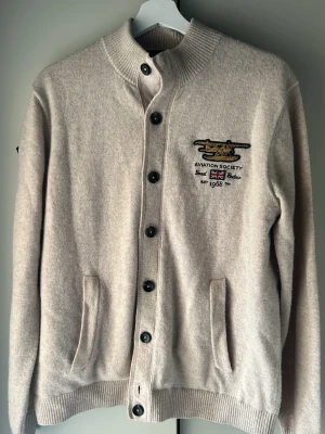 Beige cardigan från Morris med tryck - Stilren beige cardigan från Morris med knappar framtill, ribbad krage och muddar. Broderat flygplansmotiv och text på bröstet samt stort tryck på ryggen. Två fickor fram och detaljer på ärmen. Perfekt för dig som gillar unik design och en allmänt riktigt fräsch tröja . Priset inte hugget i sten