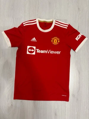 Manchester United matchtröja Adidas - Röd Manchester United matchtröja från Adidas med vita detaljer och klubbmärke på bröstet. Tröjan har korta ärmar, rund halsringning och TeamViewer-logga framtill. Tillverkad i lätt och funktionellt material, perfekt för fotbollsträning eller match.
