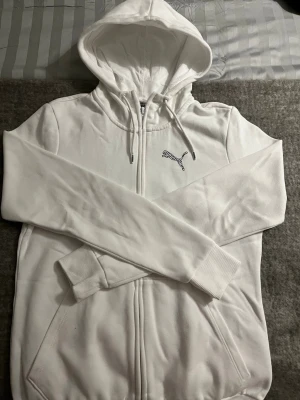 Vit zip hoodie från Puma - Säljer en clean vit hoodie från Puma med dragkedja framtill och justerbar huva. Hoodien har ribbade muddar vid ärmsluten och fickor på sidorna. Puma-logga i svart på bröstet ger en sportig vibe. Perfekt för chill dagar eller när du vill ha en enkel och snygg look.