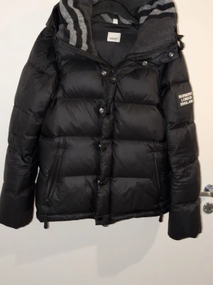 Svart dunjacka från Burberry - Snygg svart dunjacka från Burberry med huva och dragkedja. Jackan har märkets logga på ärmen och en gråmelerad insida på huvan med ränder. Perfekt för kalla vinterdagar och riktigt stilren look.