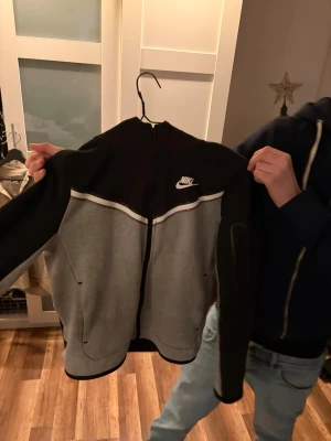 Nike svart och grå hoodie - Snygg hoodie från Nike i svart och grått med dragkedja framtill och vit Nike-logga på bröstet. Hoodien har huva och två fickor på sidorna. Tillverkad i mjukt sweatshirtmaterial som passar perfekt till en avslappnad stil. (Storlek 147-158)