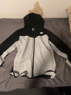 Nike Tech Fleece hoodie svart/grå - Nike Tech Fleece hoodie i svart och ljusgrått med dragkedja framtill och huva. Snygg tvåfärgad design med Nike-logga på bröstet. Tillverkad i mjukt och lätt material som passar perfekt till streetwear-stilen. Fickor på sidorna och ribbade muddar.