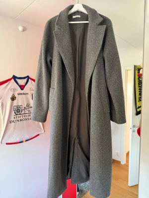 Grå lång kappa med skärp - Stilren grå kappa i klassisk modell med breda slag och knytskärp i midjan. Kappan är helfodrad och har två sidfickor. Knappt använd och i mycket bra skick!