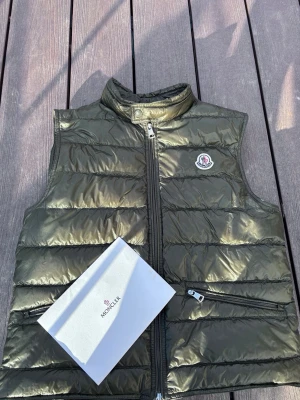Olivgrön dunväst från Moncler - Snygg olivgrön dunväst från Moncler ( ÄKTA )med hög krage och dragkedja framtill. Västen har två sidofickor med dragkedja och Moncler-logga på bröstet. Tillverkad i glansigt material och fylld med dun för extra värme. Perfekt för lager på lager under kyliga dagar. Pris diskuteras 