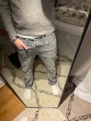 Ljusgråa jeans från Jack&jones - Säljer ett par ljusgråa jeans från jack&jones  med avslappnad passform och låg midja. Jeansen har klassiska fem fickor, vita sömmar och en svart patch med logga bak i midjan. Perfekta för en casual och trendig look.
