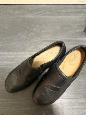Svarta loafers från Clarks - Säljer ett par svarta loafers från Clarks med rund tå och diskreta sömdetaljer. Skorna har en bekväm innersula och elastiska partier på sidorna för enkel påtagning. Yttermaterialet är slätt läder och sulan är i gummi för bra grepp.