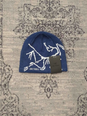 Arc'teryx mössa - Snygg blå mössa från Arc'teryx med vit grafisk dinosaurie-logga och broderad text framtill. Tillverkad i mjukt stickat material som sitter skönt på huvudet. Perfekt för dig som vill ha en stilren och sportig look. Gjord i ull