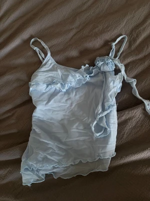 Ljusblå volang topp med knytband - Söt ljusblå linnetopp med tunna axelband, volangdetaljer längs halsringningen och asymmetrisk nederkant. Toppen har ett dekorativt knytband på ena axeln och är tillverkad i ett tunt, mjukt material som känns luftigt och somrigt.