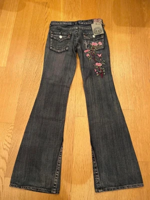 Vintage rare true religion jeans med broderi  - Snygga mörkblå bootcut jeans från True religion med broderade rosa blommor och fjärilar på ena bakfickan. Jeansen har klassisk femficksmodell, låg midja och utsvängda ben. Materialet är denim i bomull med en mjuk tvättad känsla. Midjemått ca : 37cm
