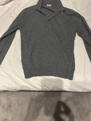 Grå stickad tröja från Rose & Born - Snygg grå stickad tröja från Rose & Born med klassisk krage och lång ärm. Tröjan har ribbade muddar vid ärmslut och nederkant, perfekt för en stilren och avslappnad look. Materialet känns mjukt och håller värmen under kyliga dagar.