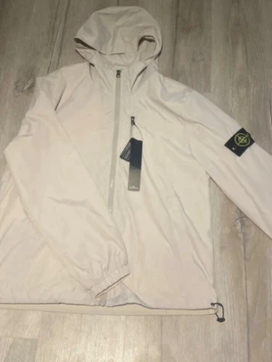 Beige vindjacka från Stone Island - Stilren beige vindjacka från Stone Island med huva och dragkedja framtill. Jackan har den klassiska logotypen på ärmen och justerbar dragsko nedtill. Perfekt för blåsiga dagar och enkel att matcha med olika outfits.