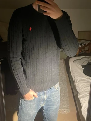 Svart kabelstickad tröja Polo Ralph Lauren - Kabelstickad svart tröja från Polo Ralph Lauren med röd broderad logga på bröstet. Tröjan har rund halsringning, långa ärmar och ribbade muddar. Klassisk och stilren design som passar till det mesta. Materialet är mjukt och bekvämt. Den har tyvärr krympt i tvätten så nu är det en m. Killen på bilden är 184 och 75kg