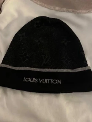 Svart mössa från Louis Vuitton allt som finns med i bilden ingår  - Snygg svart mössa från Louis Vuitton med diskret monogrammönster och grå kant. Märkets logga är broderad framtill. Tillverkad i mjukt material som känns lyxigt och bekvämt. Perfekt accessoar för dig som vill ha en exklusiv touch på din outfit. Pris kan diskuteras 